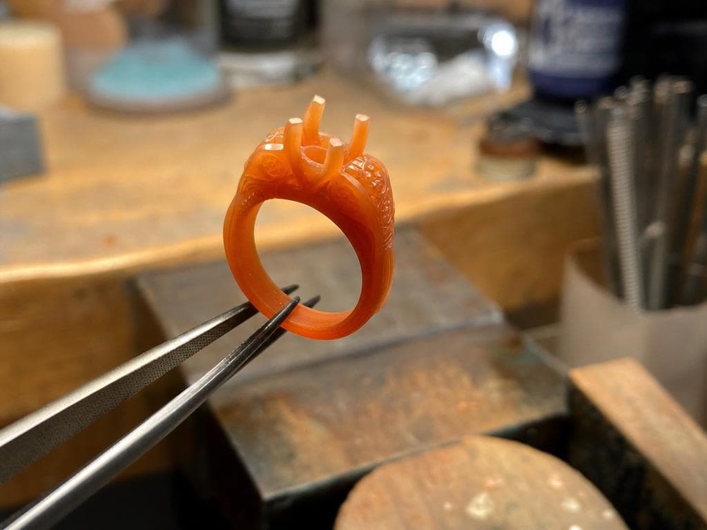 A carved orange wax prototype of the ring held in tweezers at the bench